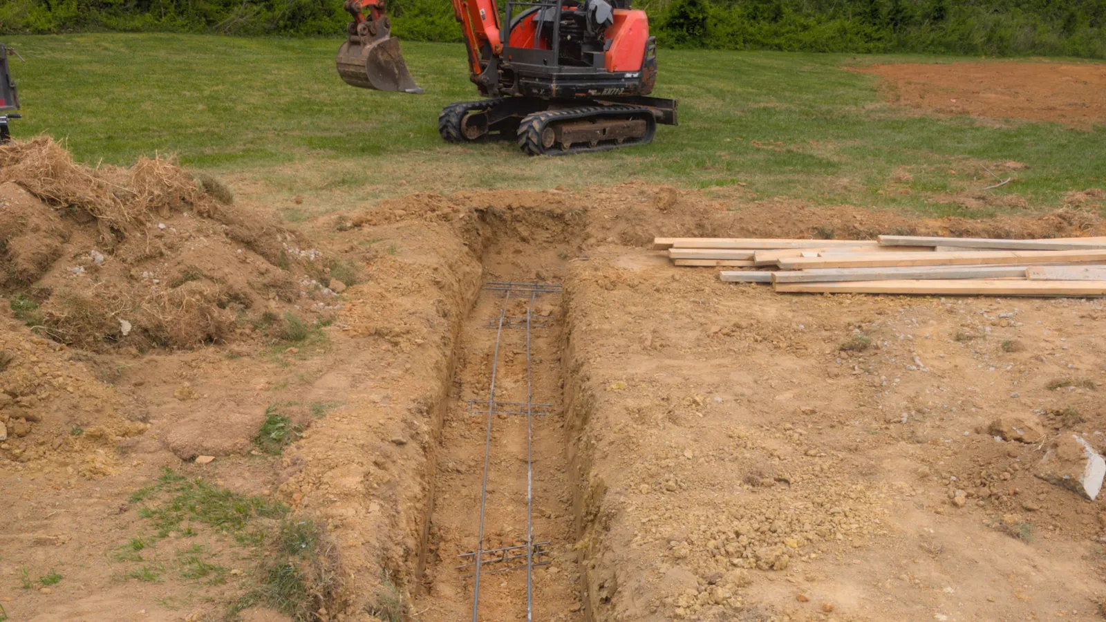Footing trench dug with rebar in place and a mini excavator behind it, ready for a concrete foundation pour