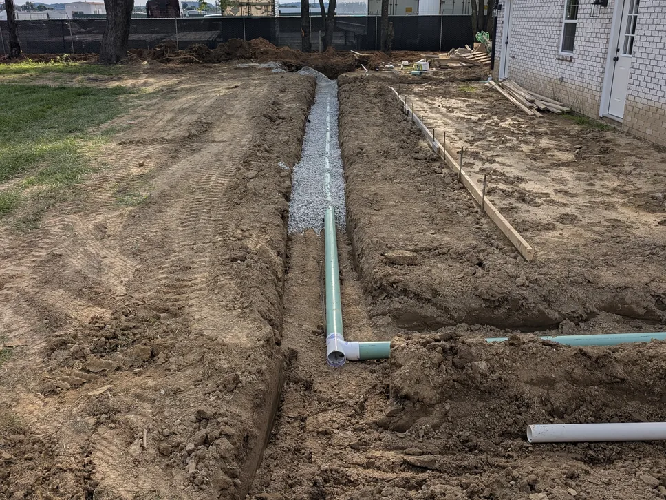 Graded yard with a drain line in a gravel trench and forms set beside a brick house, site grading